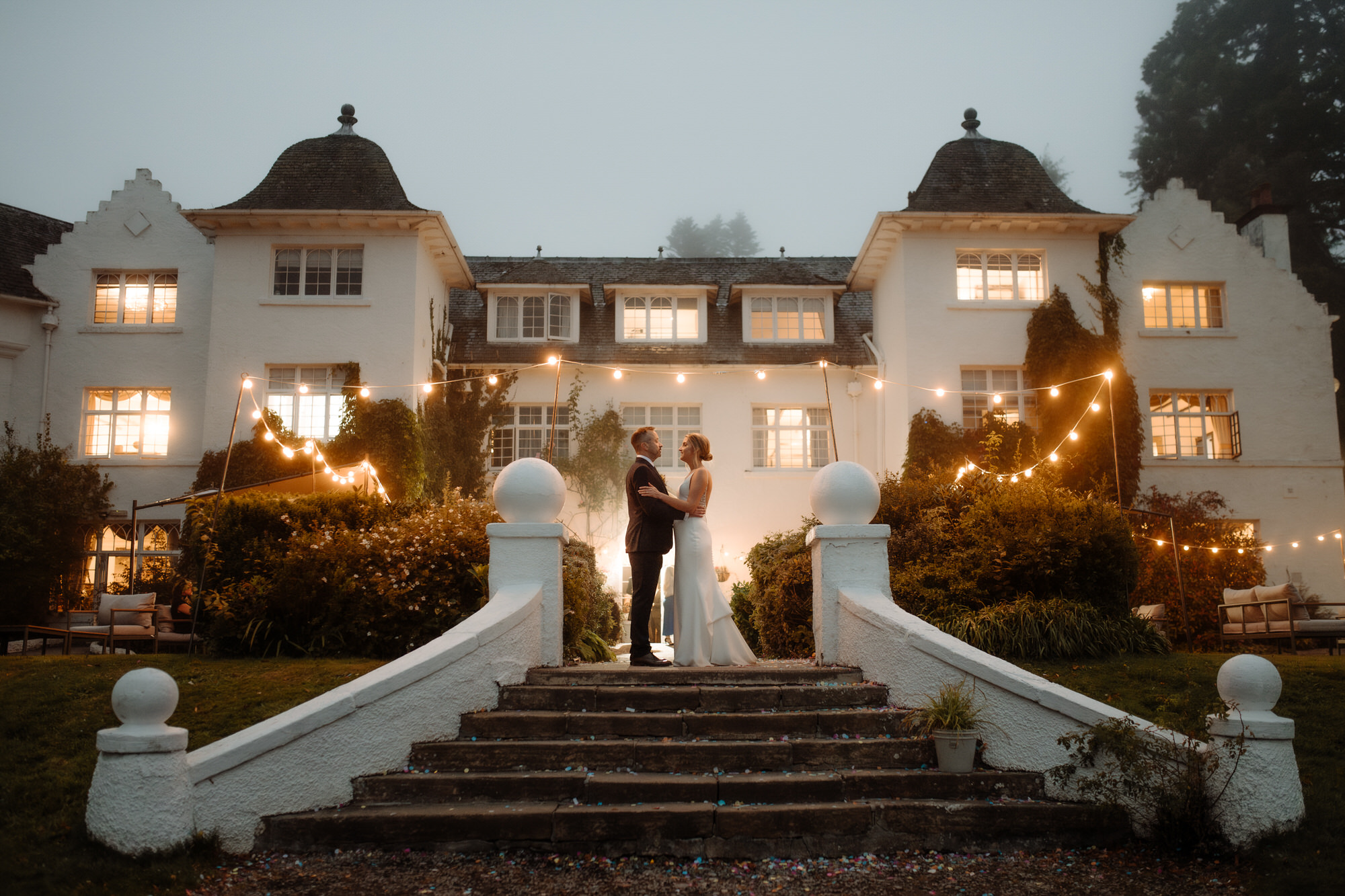 Bride and Groom stand under festoon lights at twilight at Achnagairn Castle.