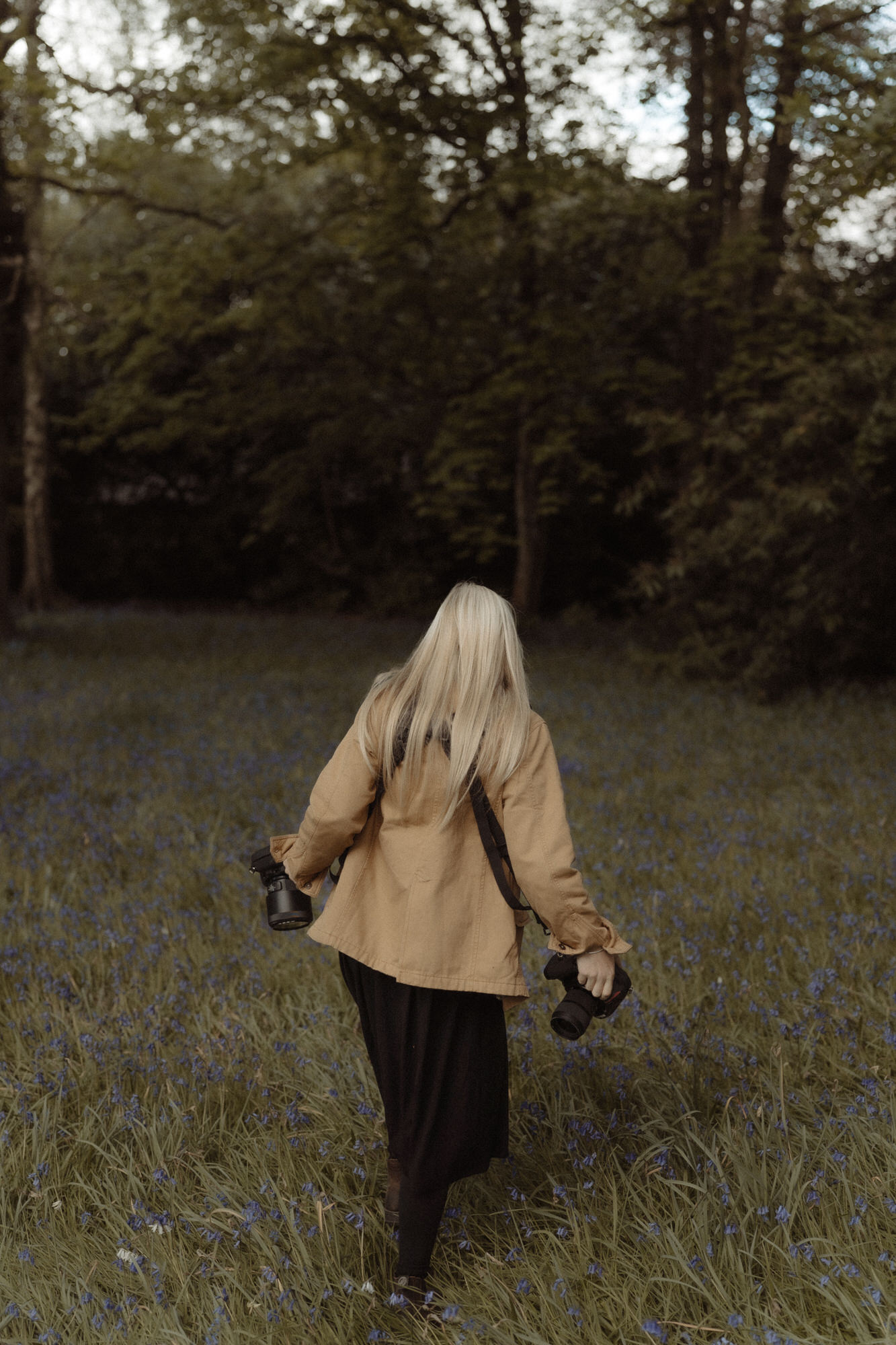 Girl in yellow jacket with cameras walks away through a bluebell meadow.