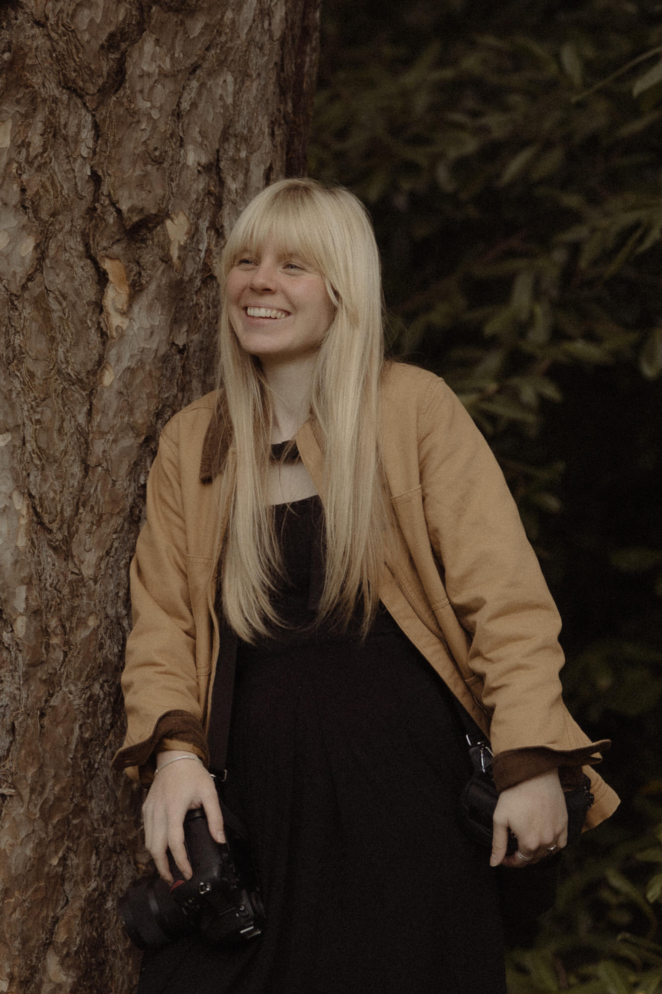 Blonde girl in yellow jacket stands beside a tree with two cameras.